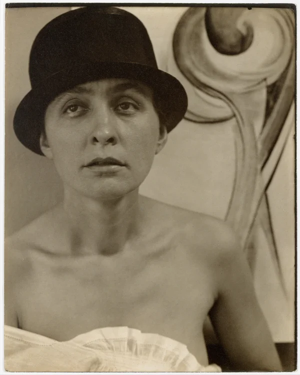 georgia-o-keeffe frontal-portrait-torso-free-shoulders-with-hat-and-painting-in-background female